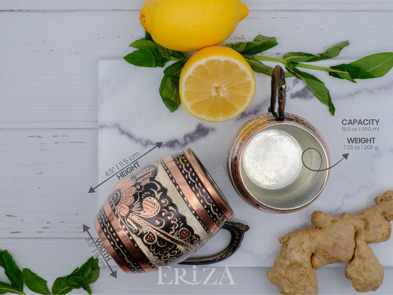 May include: Two copper mugs with intricate designs. The mug on the left has a black and white floral pattern with copper accents. The mug on the right is plain copper. Both mugs have a capacity of 18.5 ounces or 550 milliliters and weigh 7.23 ounces or 205 grams.
