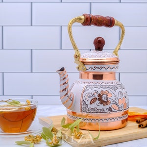May include: White enamel tea kettle with copper accents and floral designs. The kettle has a wooden handle and a gold spout. It is sitting on a wooden cutting board with cinnamon sticks and dried orange slices.