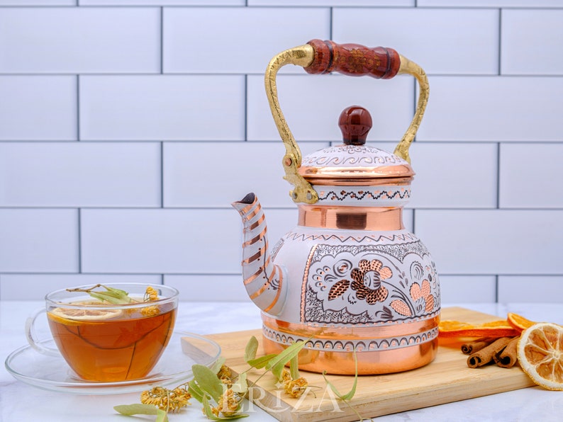 May include: White enamel tea kettle with copper accents and floral designs. The kettle has a wooden handle and a gold spout. It is sitting on a wooden cutting board with cinnamon sticks and dried orange slices.