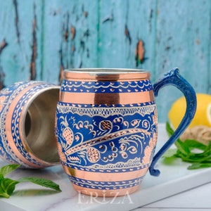 May include: Two copper mugs with blue and white floral designs and copper accents. The mug on the right has a blue handle. The mugs are on a white surface with a blue background.