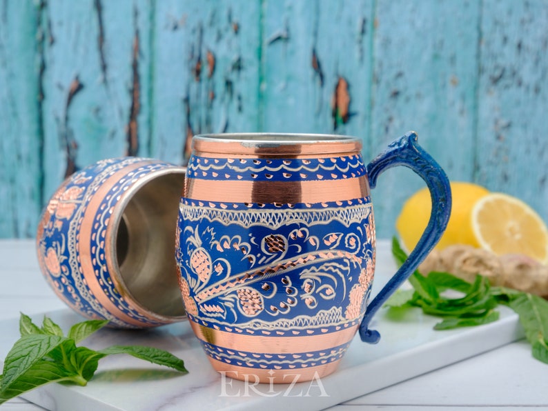May include: Two copper mugs with blue and white floral designs and copper accents. The mug on the right has a blue handle. The mugs are on a white surface with a blue background.