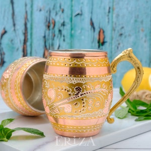 May include: Two copper mugs with ornate gold designs. The mug on the right has a gold handle. The mugs are on a white surface with green mint leaves and a lemon.