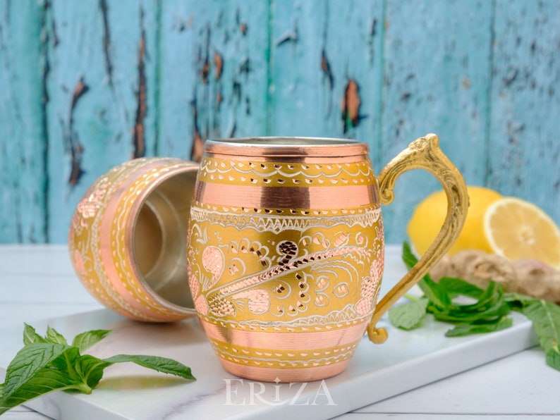 May include: Two copper mugs with ornate gold designs. The mug on the right has a gold handle. The mugs are on a white surface with green mint leaves and a lemon.
