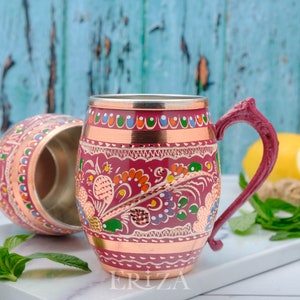 May include: Two copper mugs with colorful floral designs and a copper rim. The mug on the right has a handle. The mugs are on a white surface with green leaves and a lemon.