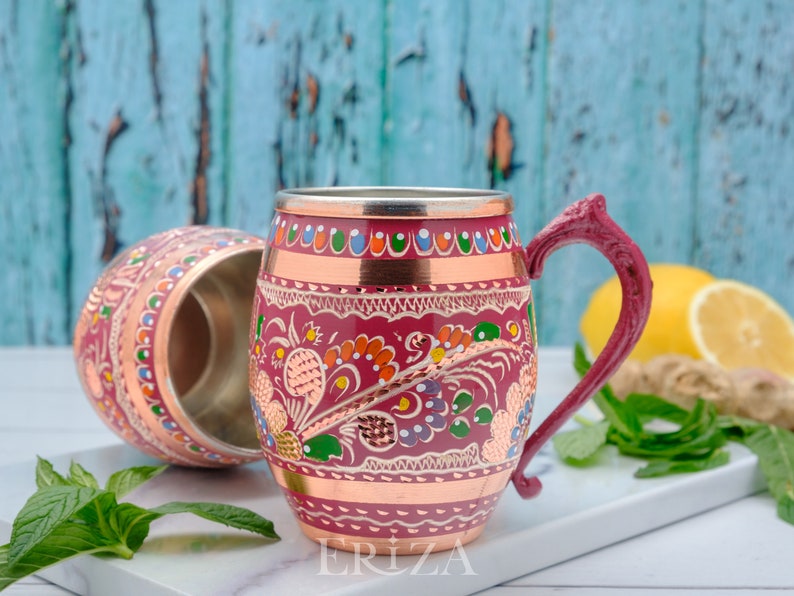 May include: Two copper mugs with colorful floral designs and a copper rim. The mug on the right has a handle. The mugs are on a white surface with green leaves and a lemon.