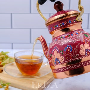 May include: A red copper teapot with a gold handle and intricate floral designs in shades of pink, blue, and green. The teapot is pouring tea into a clear glass cup.