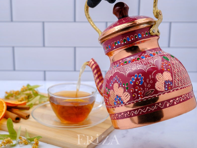 May include: A red copper teapot with a gold handle and intricate floral designs in shades of pink, blue, and green. The teapot is pouring tea into a clear glass cup.