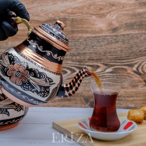 Puede incluir: Una tetera turca de cobre y negra con intrincados dise&ntilde;os florales est&aacute; vertiendo t&eacute; en un vaso de t&eacute; turco tradicional. La tetera tiene un asa rayada de cobre y negro. El vaso de t&eacute; est&aacute; sobre un platillo blanco con un dise&ntilde;o rojo y blanco. Hay baklava en una tabla de cortar de madera en el fondo.
