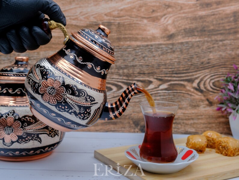 Puede incluir: Una tetera turca de cobre y negra con intrincados dise&ntilde;os florales est&aacute; vertiendo t&eacute; en un vaso de t&eacute; turco tradicional. La tetera tiene un asa rayada de cobre y negro. El vaso de t&eacute; est&aacute; sobre un platillo blanco con un dise&ntilde;o rojo y blanco. Hay baklava en una tabla de cortar de madera en el fondo.