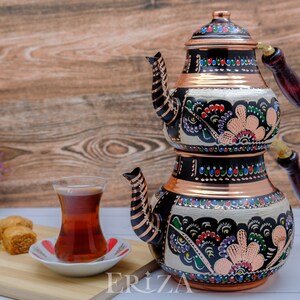 Puede incluir: Un juego de t&eacute; turco tradicional con base de cobre y dise&ntilde;o floral en blanco y negro. El juego incluye una tetera y una olla m&aacute;s peque&ntilde;a para preparar t&eacute; fuerte. La tetera tiene un asa de madera y un pico. El juego est&aacute; sobre una superficie de madera con un vaso de t&eacute; y delicias turcas.