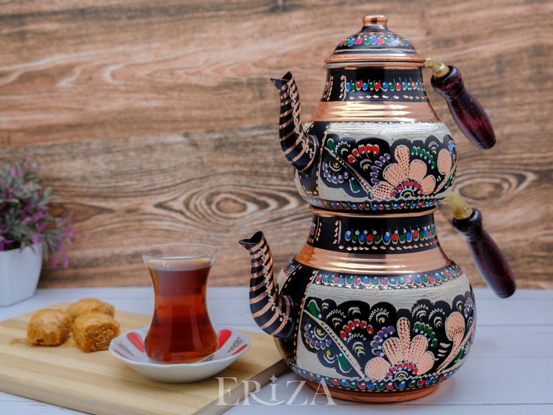 Puede incluir: Un juego de t&eacute; turco tradicional con base de cobre y dise&ntilde;o floral en blanco y negro. El juego incluye una tetera y una olla m&aacute;s peque&ntilde;a para preparar t&eacute; fuerte. La tetera tiene un asa de madera y un pico. El juego est&aacute; sobre una superficie de madera con un vaso de t&eacute; y delicias turcas.