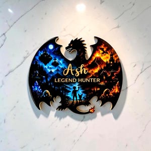 May include: A dragon-shaped wall plaque featuring a split design with a blue moonlit landscape on the left and a fiery orange volcanic scene on the right. The text Ash LEGEND HUNTER is printed in the centre above a silhouette of a person and creature.