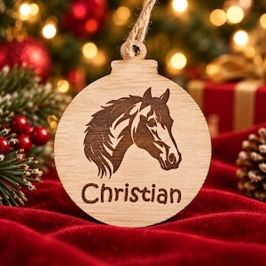 May include: A round wooden ornament featuring a laser-engraved horse head profile and the name Christian printed in a dark font. The ornament has a small hole at the top with a piece of twine attached for hanging, showcasing a natural wood grain.