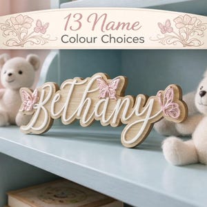 Puede incluir: Letrero de nombre de madera con el nombre "Bethany" en escritura blanca, adornado con mariposas rosas. El letrero es marrón claro con un contorno blanco. El texto "13 Name Colour Choices" se muestra arriba.