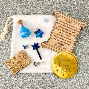 Puede incluir: Una colección de artículos con temática de hadas sobre una bolsa de tela blanca. Incluye una pequeña botella de vidrio con purpurina azul, un dije de estrella azul, un mapa de madera, una varita, una llave, un pergamino con texto, una moneda de oro con texto y una mariposa.