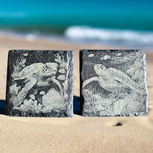 May include: Two black slate coasters with white engraved images of sea turtles swimming in a coral reef. The coasters are on a sandy beach.