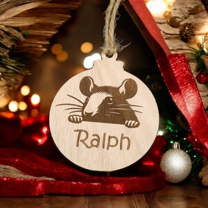 Könnte beinhalten: Ein hölzerner Weihnachtsbaumschmuck in Form einer Kugel mit einer Maus, die über den Rand schaut. Der Schmuck ist hellbraun und hat den Namen "Ralph" unten eingraviert.