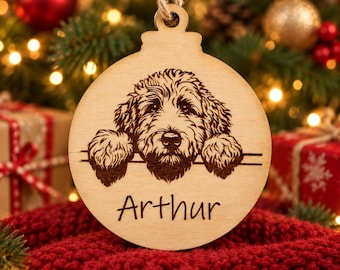May include: A round, light-colored wooden Christmas ornament featuring a laser-engraved illustration of a shaggy dog resting its paws on a horizontal line. The name Arthur is engraved in a simple script font below the dog illustration.