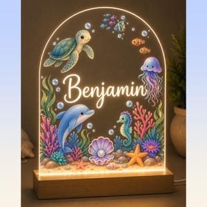 May include: An arched acrylic night light with a wooden base featuring a colourful underwater scene with a sea turtle, dolphin, seahorse, jellyfish, and clownfish. The name Benjamin is printed in white script in the centre of the display.