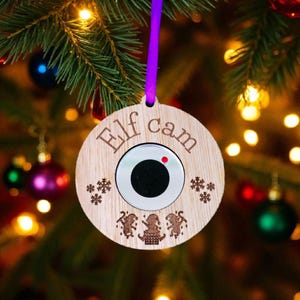 May include: A wooden Christmas ornament with the words "Elf cam" engraved on it. The ornament has a circular camera lens in the center and is decorated with snowflakes and elf figures. A purple ribbon is attached for hanging.