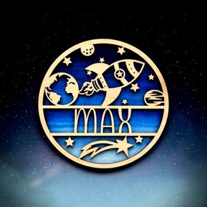 Puede incluir: Un letrero de pared circular de madera con un diseño recortado de un cohete, el planeta Tierra, estrellas y un cometa. El nombre MAX se muestra en el centro en letras negritas y claras sobre un fondo azul con degradado de color.