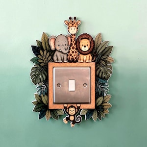 May include: A light switch cover plate featuring a wooden frame surrounded by illustrations of a grey elephant, a spotted giraffe, a brown lion, and a hanging monkey, all nestled among green tropical leaves on a light green wall background.