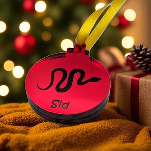 May include: A red Christmas bauble featuring a black snake design and the name "Sid". The bauble has a black border and a yellow ribbon. The background shows a Christmas tree with lights and a wrapped gift.