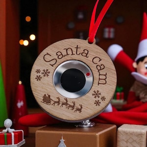 Może przedstawiać: Drewniana ozdoba świąteczna z wygrawerowanym napisem "Santa cam". Ozdoba ma srebrny środek i czerwoną wstążkę do zawieszenia. Wygrawerowana jest również mała ilustracja sań Świętego Mikołaja i reniferów.