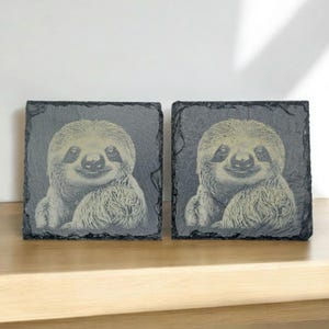 May include: Two black slate coasters featuring a white image of a sloth on each. The sloth has a smiling face and is looking directly at the viewer.