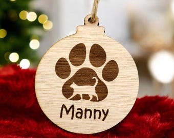 May include: A wooden Christmas ornament shaped like a ball with a paw print and a cat silhouette inside. The ornament is personalized with the name "Manny".