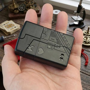 Könnte beinhalten: Ein schwarzes, rechteckiges Puzzle mit Spielkartensymbolen und geometrischen Mustern. Das Puzzle wird in einer Hand gehalten, mit einem roten Beutel und anderen Objekten im Hintergrund.