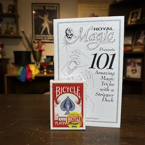 May include: A Bicycle Stripper Deck of playing cards with a red and white design, next to a booklet titled "Royal Magic Presents 101 Amazing Magic Tricks with a Stripper Deck". The image is set on a dark wooden surface.