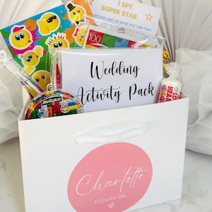 May include: A white gift bag with a pink circle logo that says "Charlotte Flower Girl" and the text "Wedding Activity Pack". The bag is filled with various items, including a sticker sheet, a game, and sweets.