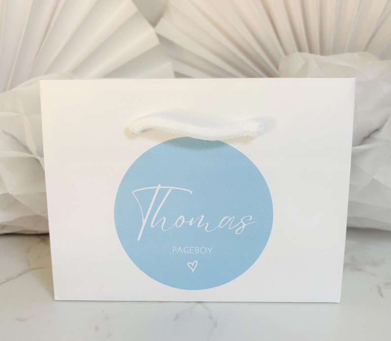 May include: White gift bag with a blue circle and the text "Thomas Pageboy" in white script. A small white heart is below the text.