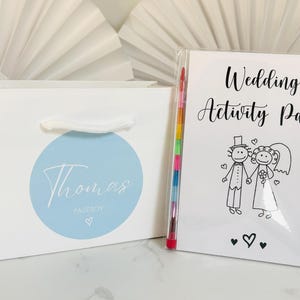 May include: A white gift bag with a blue circle and the text "Thomas Pageboy" on it. Next to the bag is a white booklet with the text "Wedding Activity Pack" and a black and white illustration of a bride and groom. A rainbow colored crayon is sticking out of the booklet.