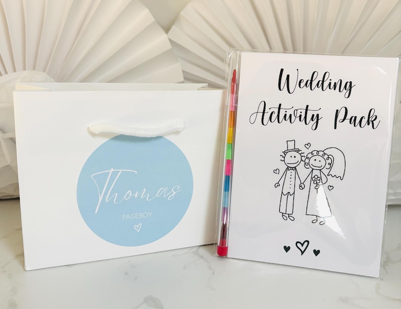 May include: A white gift bag with a blue circle and the text "Thomas Pageboy" on it. Next to the bag is a white booklet with the text "Wedding Activity Pack" and a black and white illustration of a bride and groom. A rainbow colored crayon is sticking out of the booklet.