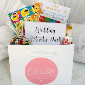 May include: A white gift bag with a pink circle sticker that says "Charlotte Flower Girl" and a heart. The bag is filled with wedding-themed activities for kids, including a "Wedding Activity Pack" card, a "I Spy Super Star" game, a smiley face sticker sheet, a small toy, and candy.