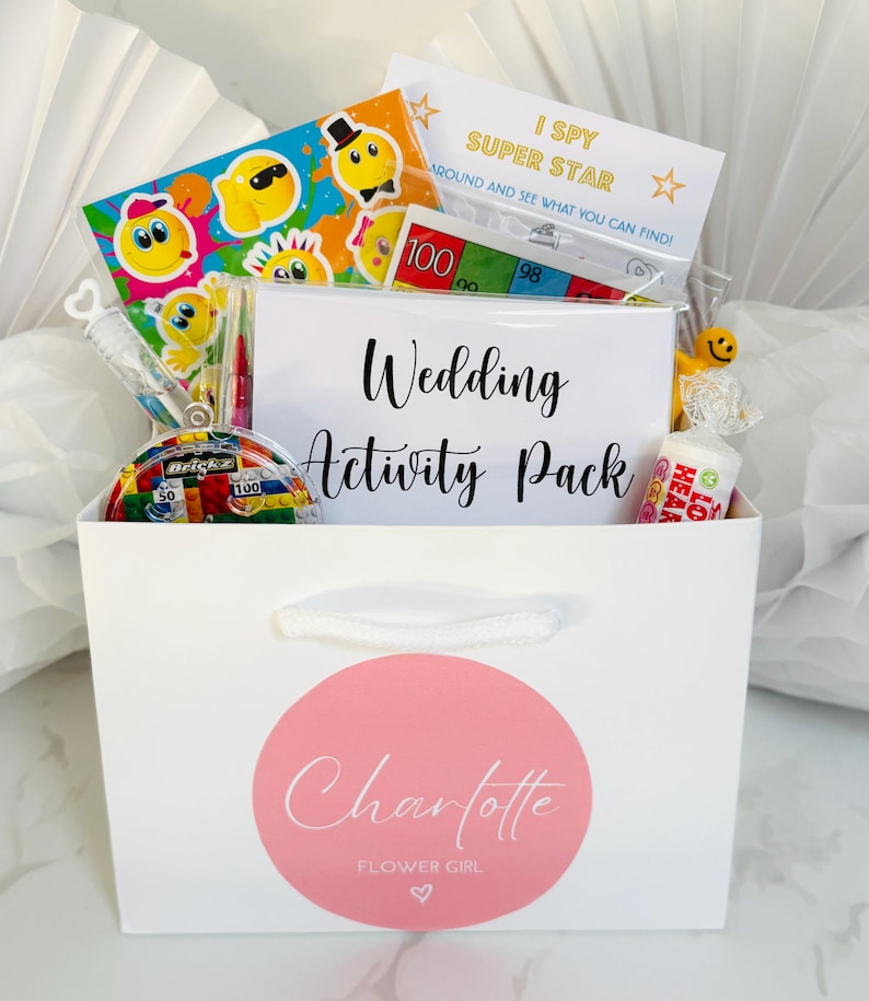 May include: A white gift bag with a pink circle sticker that says "Charlotte Flower Girl" and a heart. The bag is filled with wedding-themed activities for kids, including a "Wedding Activity Pack" card, a "I Spy Super Star" game, a smiley face sticker sheet, a small toy, and candy.