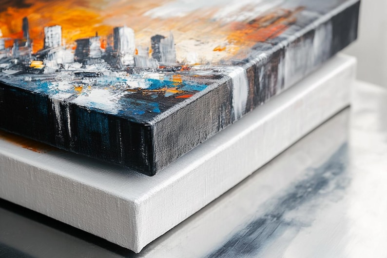 May include: Two canvases stacked on top of each other. The top canvas has an abstract cityscape painting with orange, blue, and white paint. The bottom canvas is white.
