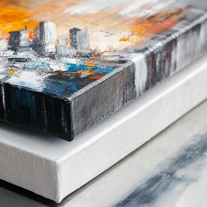 May include: Two canvases stacked on top of each other. The top canvas has an abstract cityscape painting with orange, blue, and white paint. The bottom canvas is white.