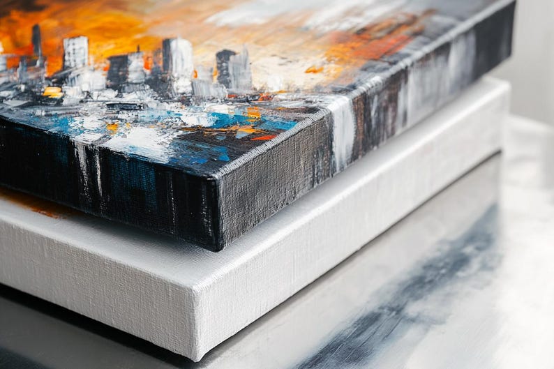 May include: Two canvases stacked on top of each other. The top canvas has an abstract cityscape painting with orange, blue, and white paint. The bottom canvas is white.
