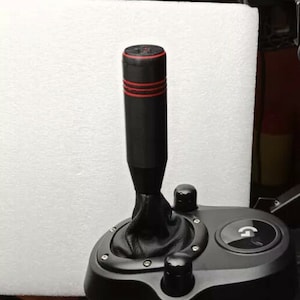 May include: Black gear shift knob with a red accent ring. The knob is attached to a black gear shift base with a leather boot.