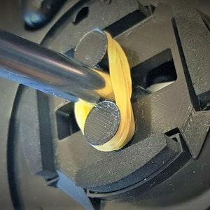 May include: Close-up of a black mechanical component with a yellow rubber band looped through it. A silver screwdriver is inserted into the mechanism. The component has a circular shape with various geometric cutouts and black cylindrical pieces.