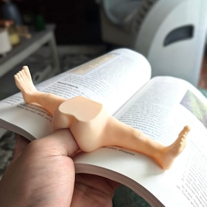 May include: A whimsical, tan-colored bookend shaped like a pair of legs, resting on the pages of an open book. The bookend is designed to appear as if a person is lounging on the book. The book is open to a recipe.