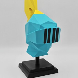 May include: A low-poly style knight helmet sculpture featuring a bright blue main section, a yellow plume on top, and a black visor with four vertical slits. The helmet is mounted on a black square base with a central support pillar.