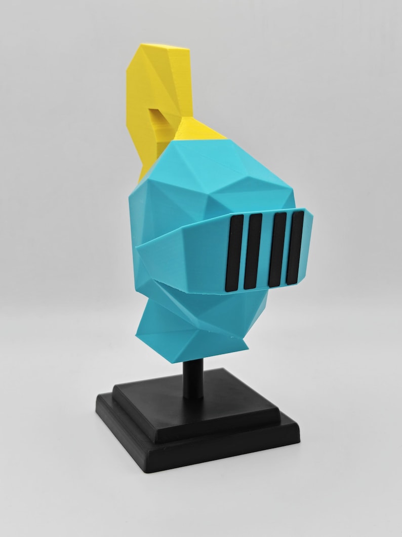 May include: A low-poly style knight helmet sculpture featuring a bright blue main section, a yellow plume on top, and a black visor with four vertical slits. The helmet is mounted on a black square base with a central support pillar.