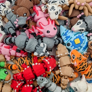 May include: A collection of colorful, articulated animal toys. The assortment includes tigers, axolotls, spiders, and other creatures in various colors like pink, red, and orange. The toys appear to be made of plastic and are designed with detailed features.