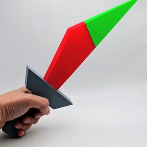 May include: A geometric toy sword with a gray handle and a blade divided into red and green sections. The toy is held in a hand against a white background, showcasing its colorful design.