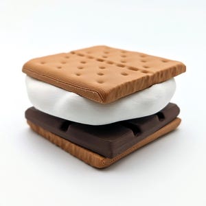 May include: A stacked s'more with a graham cracker top and bottom, a white marshmallow center, and a dark chocolate layer. The graham crackers are light brown with small dot details. The chocolate is dark brown, and the marshmallow is white.