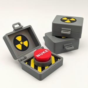 May include: A gray miniature box with a red button labeled "NUKE" and a yellow and black hazard pattern. The box is open, revealing the button. Two additional gray boxes with a yellow radiation symbol are stacked behind it.